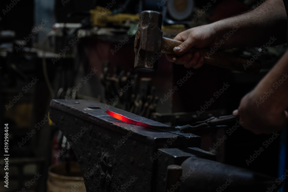 Close up view of heated metal and anvil. Blacksmith in the production process of other metal products handmade in the forge. Metalworker forging metal with a hammer into knife. Metal craft industry. Close up view of heated metal and anvil. Blacksmith in the production process of other metal products handmade in the forge. Metalworker forging metal with a hammer into knife. Metal craft industry.