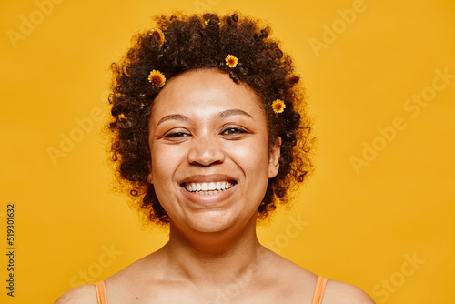 Close up portrait of happy black woman with flowers in hair smiling at camera against vibrant yellow background