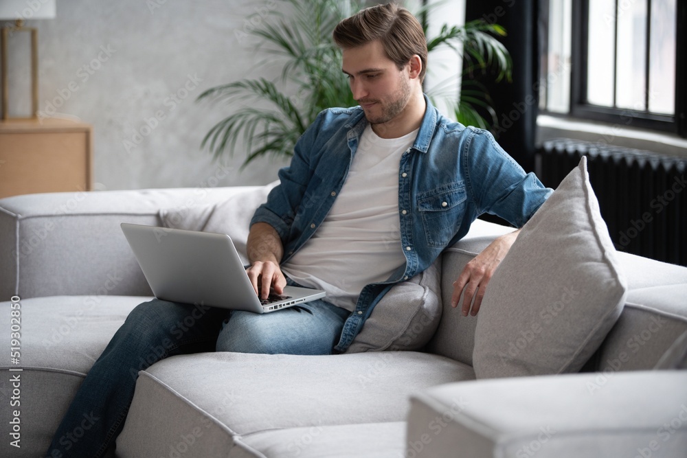 Young attractive smiling man is browsing at his laptop, sitting at home on the cozy sofa at home, wearing casual outfit. Young attractive smiling man is browsing at his laptop, sitting at home on the cozy sofa at home, wearing casual outfit.