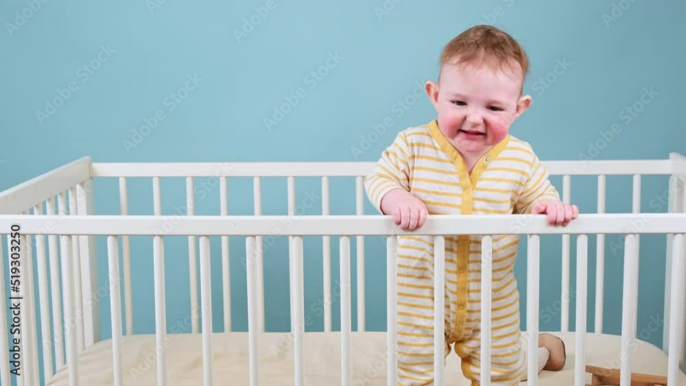 Cry infant baby boy stands in the crib, studio blue background. Sad child in yellow pajamas Cry infant baby boy stands in the crib, studio blue background. Sad child in yellow pajamas