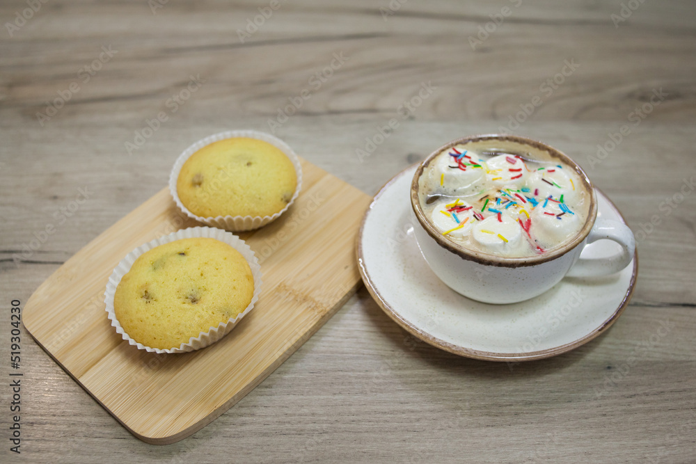 Two muffins with raisins on a wooden tray and a cup of hot chocolate with marshmallows and colored sprinkles. Two muffins with raisins on a wooden tray and a cup of hot chocolate with marshmallows and colored sprinkles.