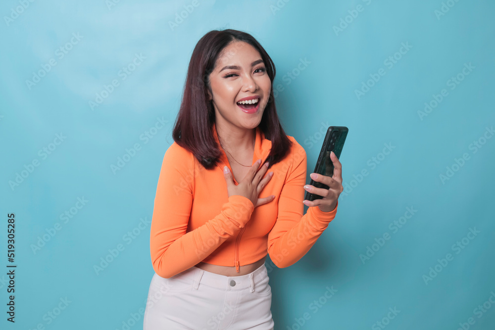 A portrait of a cheerful Asian woman looking at her smartphone over blue background