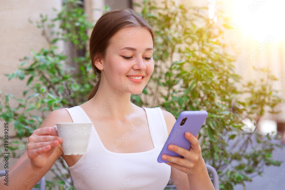 portrait of beautiful happy smiling girl, young woman sitting outdoors on terrace in cafe on table drinking beverage tea or coffee holding in hand smartphone, using looking at cell mobile phone portrait of beautiful happy smiling girl, young woman sitting outdoors on terrace in cafe on table drinking beverage tea or coffee holding in hand smartphone, using looking at cell mobile phone