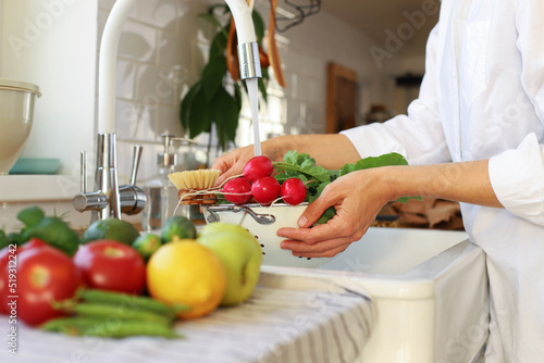A young girl washing vegetables and fruits in the kitchen in the sink.
