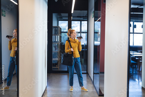 Mid adult caucasian businesswoman talking through mobile phone speaker in corridor of office