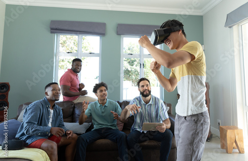 Multiracial friends looking at man wearing virtual reality simulator boxing while sitting on sofa