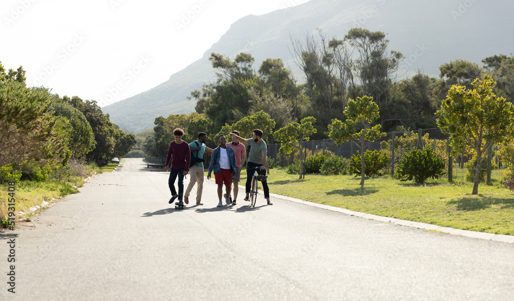 Obraz premium Male multiracial friends with bicycle walking on road amidst lush trees against clear sky in summer