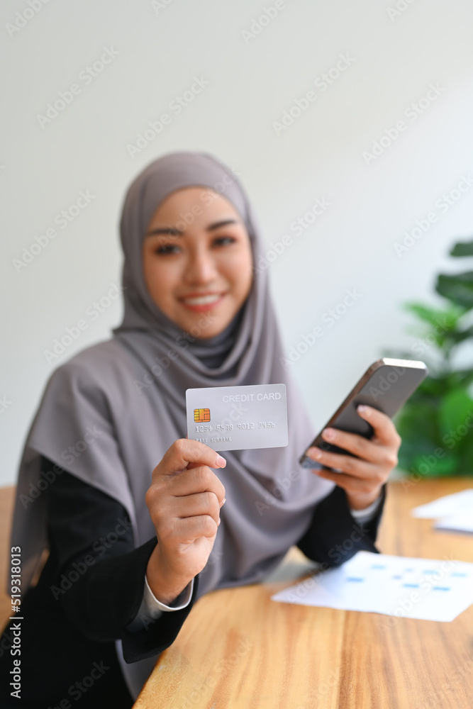 Smiling young Muslim woman holding credit card and mobile phone. Online shopping, internet banking concept Smiling young Muslim woman holding credit card and mobile phone. Online shopping, internet banking concept