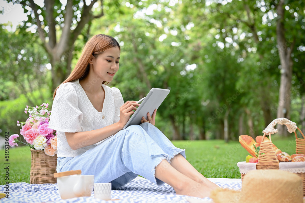 Fototapeta premium Happy young Asian female using tablet while picnicking in the green garden in summer day.