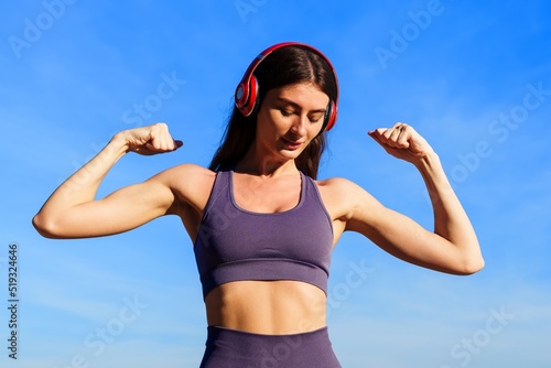 The athlete shows her biceps against the blue sky. A young woman listens to music while exercising outside in summer