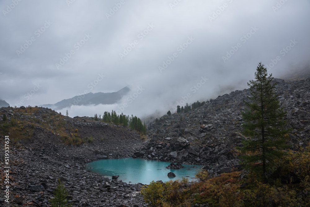Dark atmospheric landscape with turquoise alpine lake against stone hill with forest and silhouette of mountain range in dense fog. Small mountain lake and black rocks in thick low clouds during rain.
