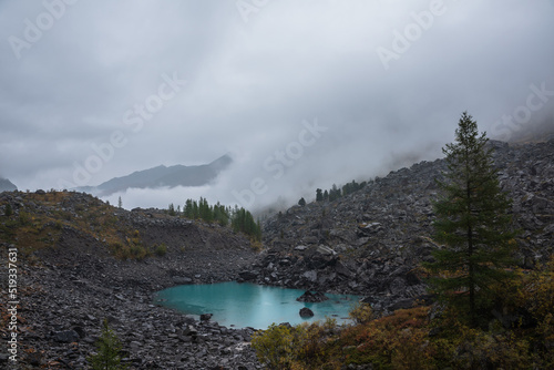 Dark atmospheric landscape with turquoise alpine lake against stone hill with forest and silhouette of mountain range in dense fog. Small mountain lake and black rocks in thick low clouds during rain.