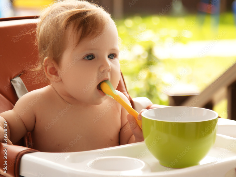 Closeup portrait of charming hungry little female girl sitting with spoon in hands and eating porridge or fruit puree, looking at camera Closeup portrait of charming hungry little female girl sitting with spoon in hands and eating porridge or fruit puree, looking at camera