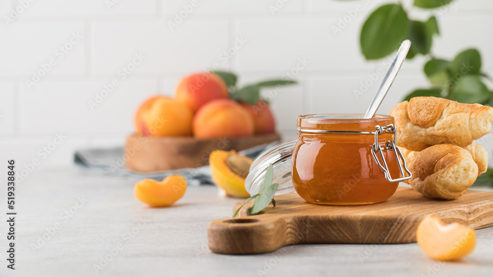 A jar of apricot jam with a spoon inside and with croissants, ripe apricots in a plate and a branch with green leaves. Fruit preservation. A jar of apricot jam with a spoon inside and with croissants, ripe apricots in a plate and a branch with green leaves. Fruit preservation.
