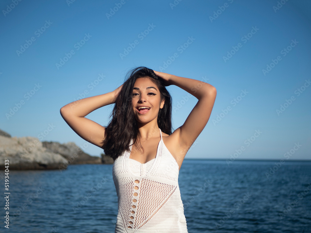 Joyful young Hispanic woman touching hair and smiling at camera near sea