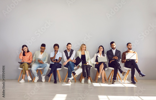 Group of multiracial people are waiting their turn for interview sitting on chairs in row. People with paper documents, resumes, laptops and mobile phones are sitting in line in personnel department.