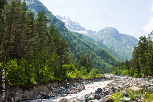 Pastoral landscape - the Terskol mountain river flows in the mountains between the slopes of high cliffs among bright green trees on a sunny summer day in the Elbrus region of Russia