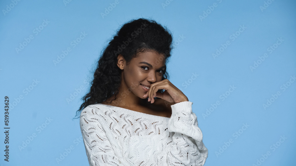 shy african american woman in white knitted sweater smiling isolated on blue. shy african american woman in white knitted sweater smiling isolated on blue.