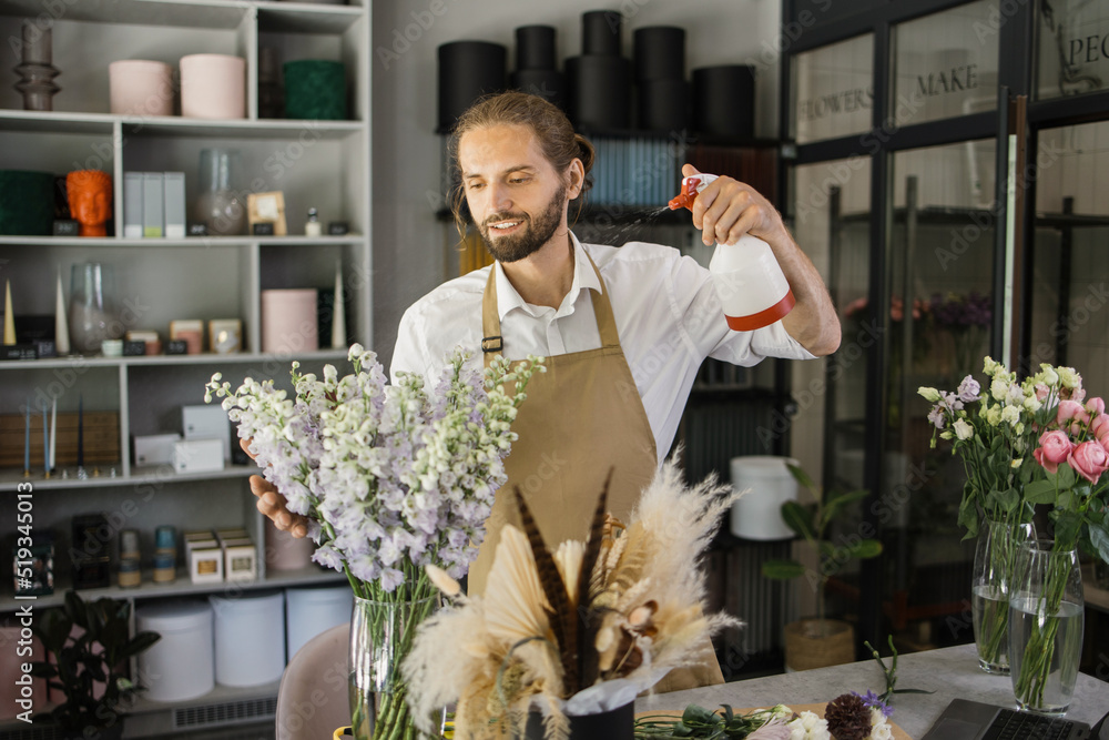 Smiling florist in uniform work in a flower shop behind counter taking care of different varieties of flowers by moisturizing them. Small business concept. Modern loft interior. Stock Photo Adobe Stoc...