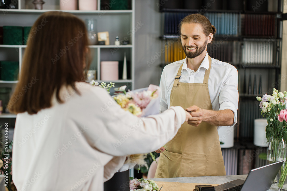 Fototapeta premium Hadsome bearded male florist receiving discount card from regular female customer and making payment at terminal in flower shop.
