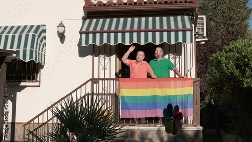 Gay couple talking while standing in the entrance of their house with a rainbow LGBT flag hanging on the railing.