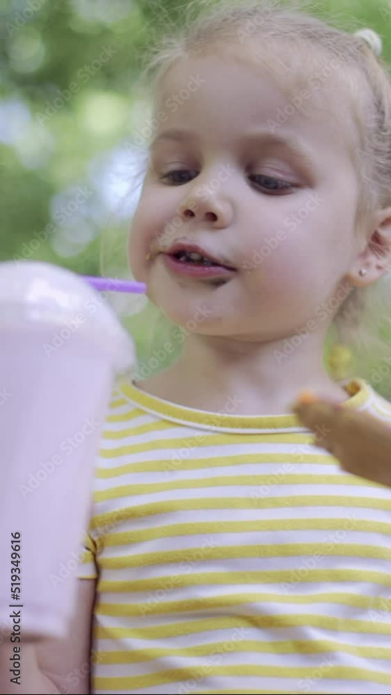 VERTICAL VIDEO: Little girl eats a colorful gingerbread and holds a milkshake in her hand. Close-up portrait of cute child girl sitting on park bench and eating cookies with a milkshake. VERTICAL VIDEO: Little girl eats a colorful gingerbread and holds a milkshake in her hand. Close-up portrait of cute child girl sitting on park bench and eating cookies with a milkshake.