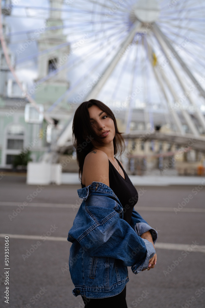 Stylish beautiful young brunette woman model in summer , dressed in a black dress and jeans jacket. Portrait, vertical photo , ferris wheel in the background. Street fashion.