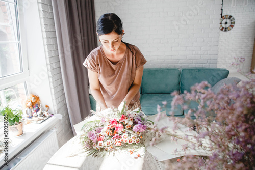A woman brunette European florist packs a multi-branch bouquet in white paper on the table in her flower studio