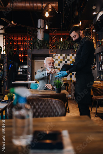 The waiter taking order for a delicious meal to the senior businessman at the restaurant. He wears a protective mask as part of security measures against the Coronavirus pandemic.