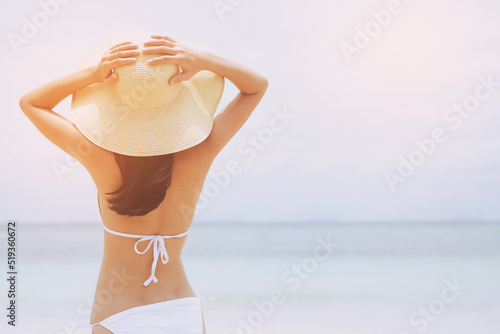 backside young woman skin tan in sunhat and bikini standing with her arms raised to her head enjoying on the beach vacation travel. and enjoy life at sea looking view of beach ocean on hot summer day.