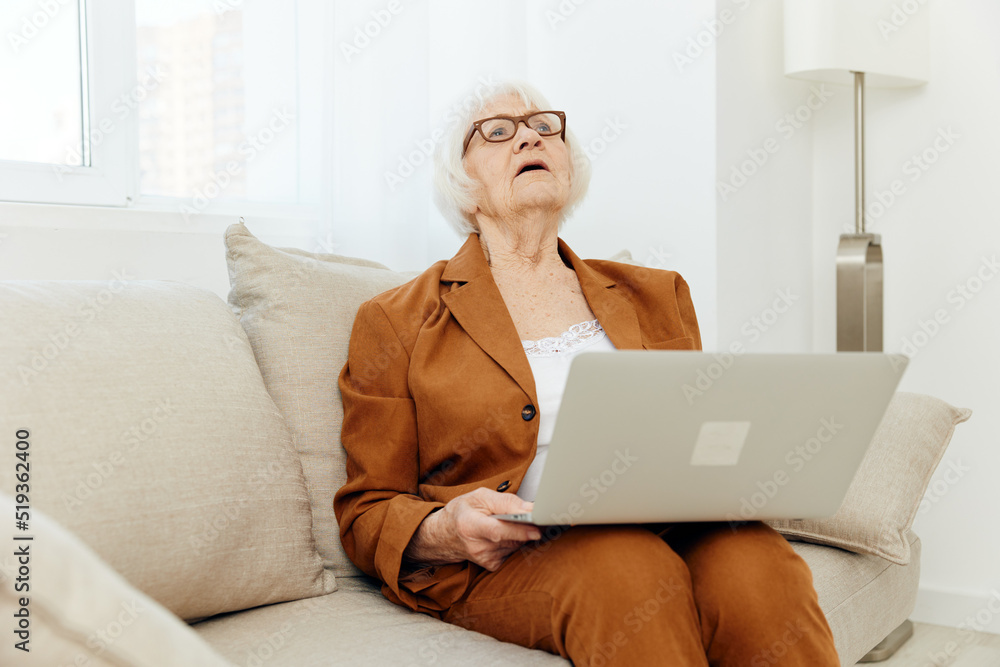 a sad elderly woman with gray hair is sitting on the sofa in the apartment holding a laptop on her lap and tired takes her head away from the monitor closing her eyes a sad elderly woman with gray hair is sitting on the sofa in the apartment holding a laptop on her lap and tired takes her head away from the monitor closing her eyes