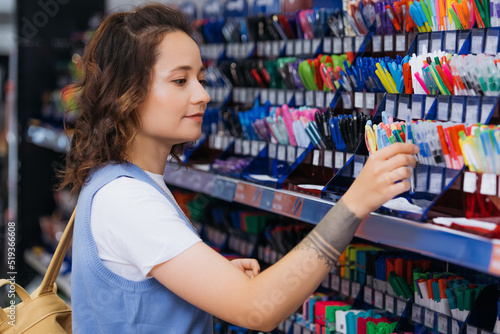 young tattooed woman near rack with multicolored pens in stationery store.