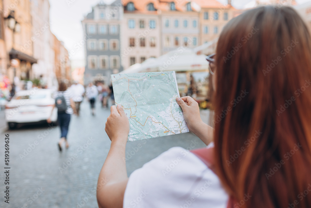Attractive young female tourist is exploring new city. Redhead girl walking in the city and holding a paper map in a warm sunny day. Urban lifestyle concept. Traveler