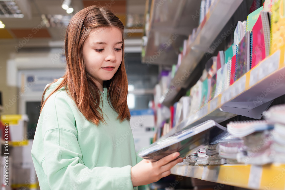 schoolgirl looking at new copybook near blurred rack in stationery shop.