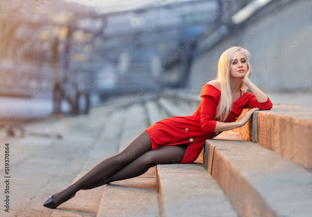 Beautiful girl in red dress with perfect legs in pantyhose and shoes with  high heels sitting on the street. Stock Photo