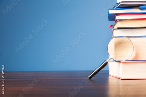 Stack book with magnifying glass on wooden desk in information library of school or university  concept for education  reading  study  copy space on blue background.
