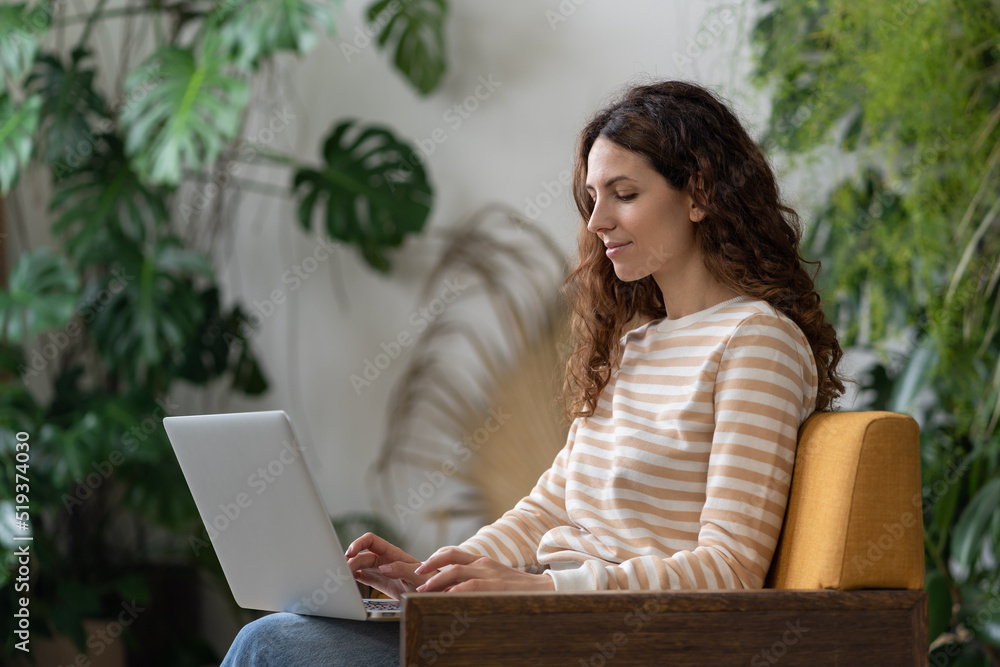 Freelancer woman sitting in armchair in green house, working on laptop surrounded by houseplant. Home garden, remote work, distance job. Female typing text on computer in cozy apartment with plants. Freelancer woman sitting in armchair in green house, working on laptop surrounded by houseplant. Home garden, remote work, distance job. Female typing text on computer in cozy apartment with plants.