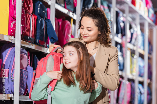 smiling woman trying new backpack on daughter in stationery store.