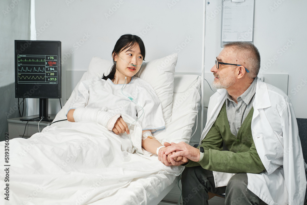 Mature man visiting his daughter in hospital sitting on chair holding her hand and talking about something Mature man visiting his daughter in hospital sitting on chair holding her hand and talking about something