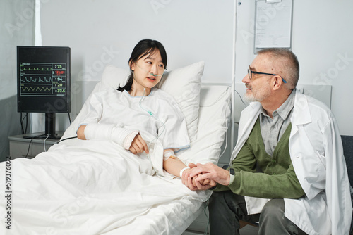 Mature man visiting his daughter in hospital sitting on chair holding her hand and talking about something