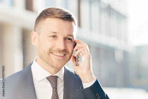 Confident happy middle aged businessman communicates via cell phone, discusses financial report with business partner, wears formal clothes, stands outdoor against blurred background. Busy CEO photo