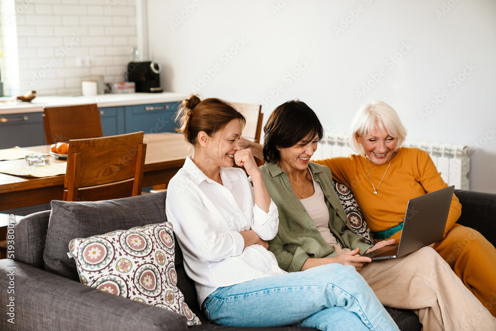 Mature three women using laptop and laughing while sitting on sofa