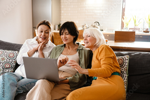 Mature three women using laptop and smiling while sitting on sofa