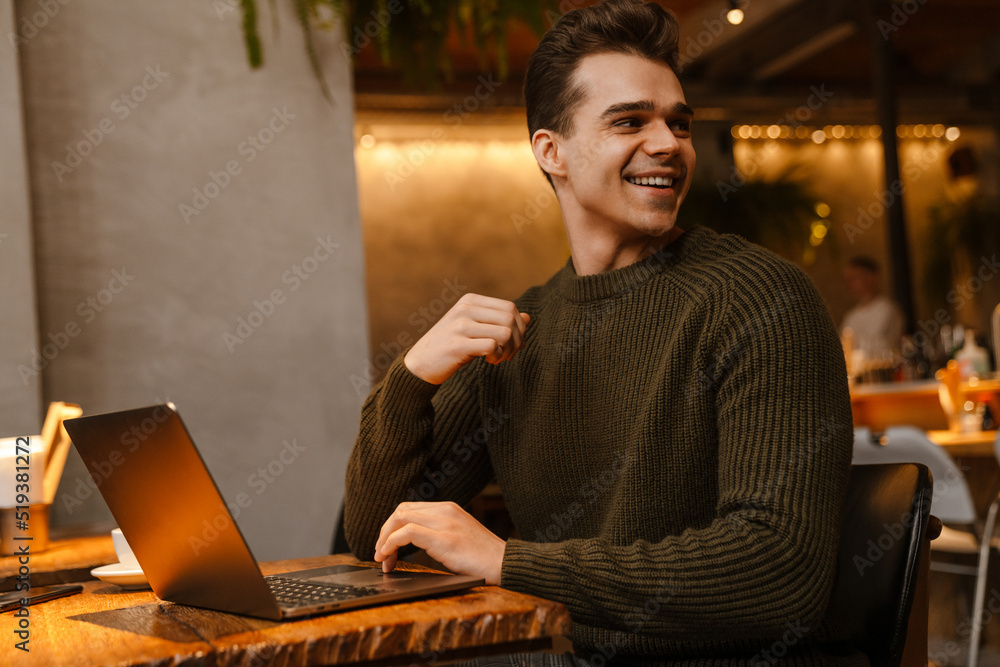 Young man smiling and looking backward while using laptop in cafe Young man smiling and looking backward while using laptop in cafe
