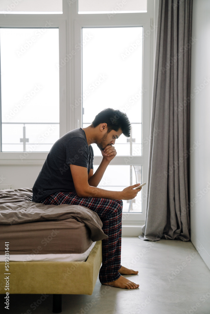 Young handsome indian man sitting on bed with phone Young handsome indian man sitting on bed with phone
