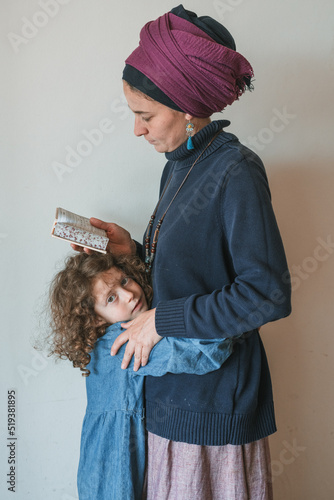 Young jewish woman with a covered head prays with a siddur (jewish prayer book) in her hands. Her little daughter hugs her tightly (63)