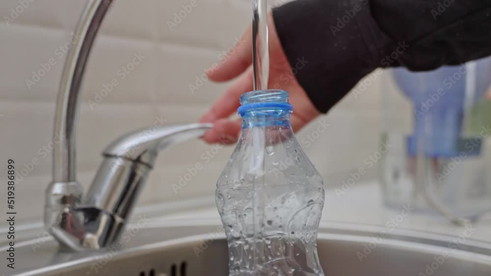 Close-up of a man in a black jacket pouring cold tap water into a plastic bottle in a home sink in the kitchen against a white wall. Close-up of a man in a black jacket pouring cold tap water into a plastic bottle in a home sink in the kitchen against a white wall.