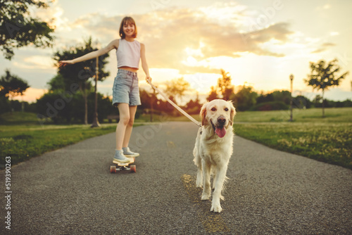 Portrait of teenage girl petting golden retriever outside in sunset running on her longboard with dog