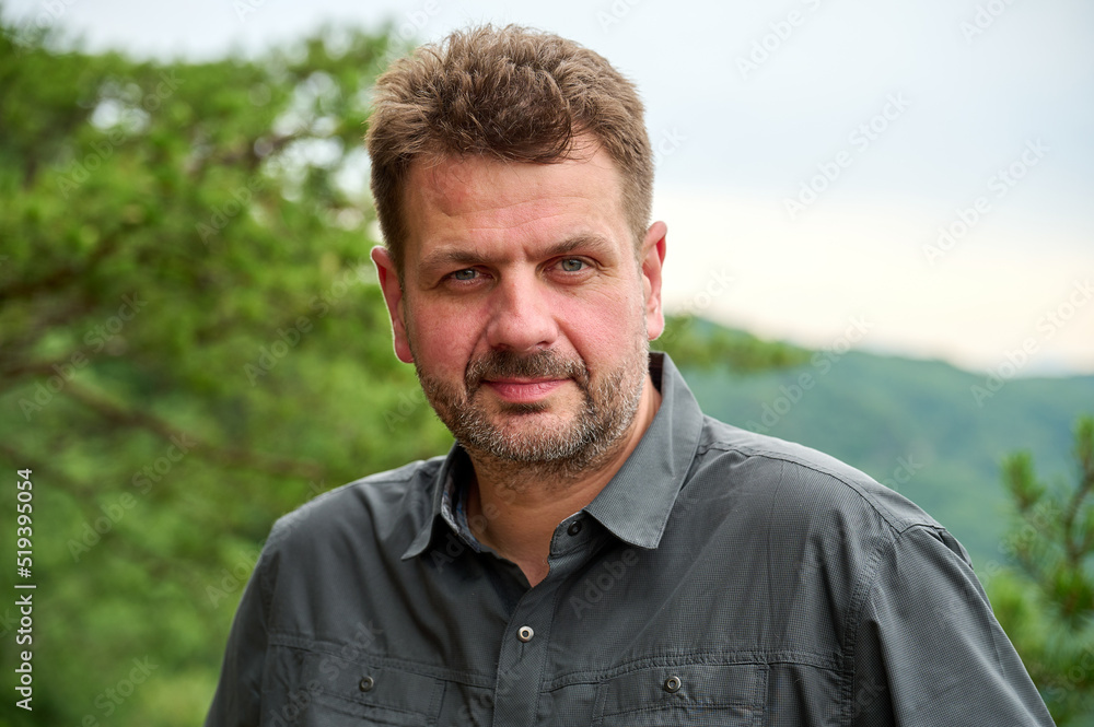 a middle-aged white caucasian man with a short haircut is looking at you with his squinted eyes and a serious facial expression. he is outdoors, with pine tree branches in the background. 