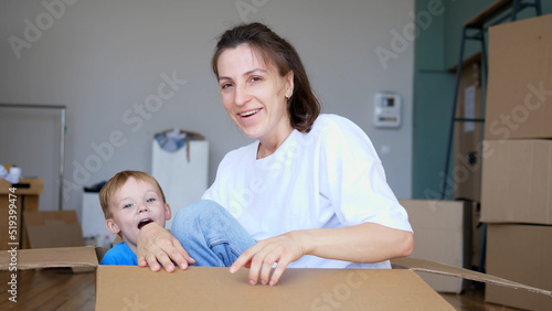 Young mother and a little son are having fun in a new house, jumping out of a cardboard box. Moving, renovation, new life.